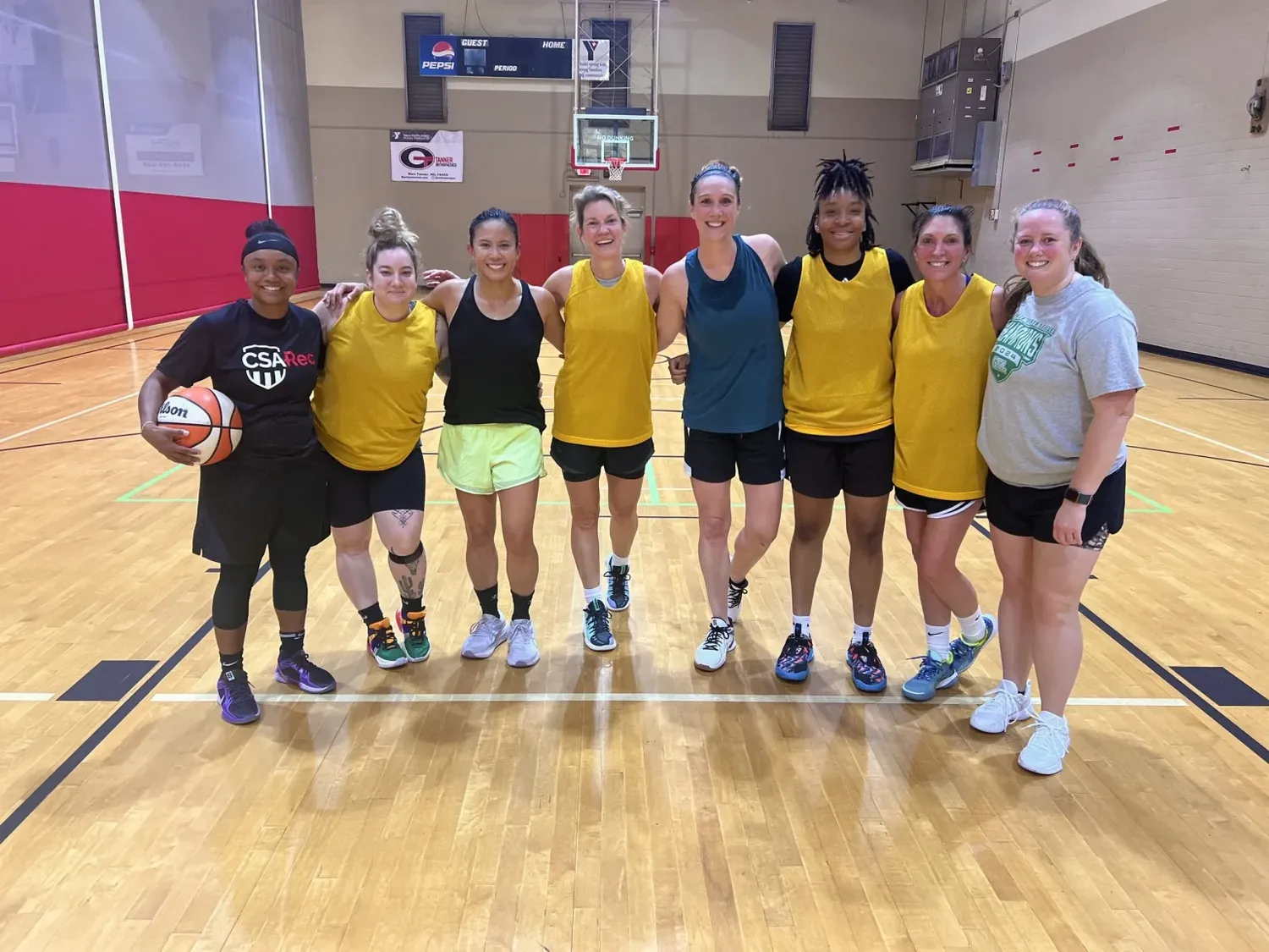 Eight women in yellow and dark jerseys stand arm in arm on an indoor basketball court, one holding a ball.